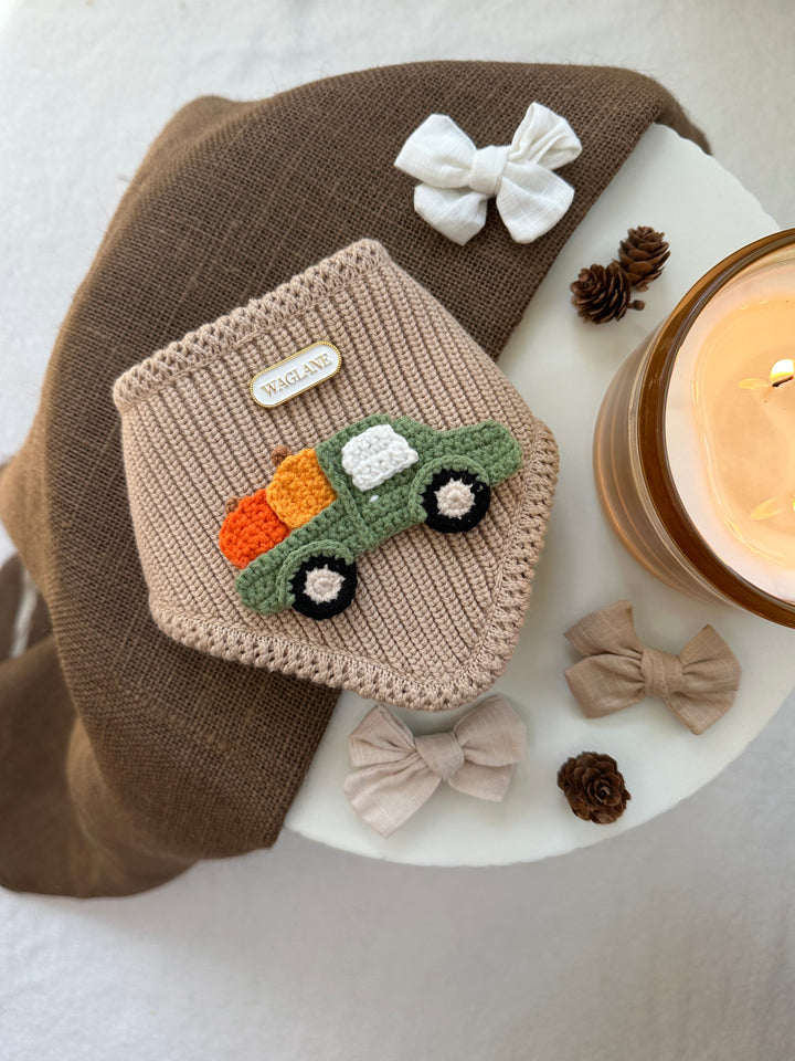 Knitted dog  bandana with crochet applique, surrounded by small bows and a candle on a textured round surface.