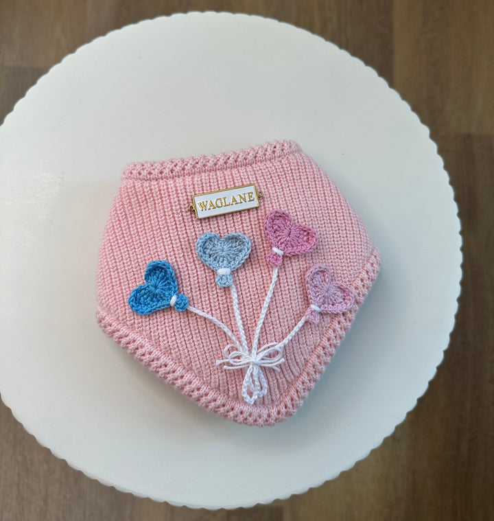Pink knit dog bandana with crochet heart-shaped balloon appliqués in blue, lilac, and rose tones, tied with a white yarn string tied in a bow and finished with a white and gold rectangular Waglane™ logo all placed on a round white platform.