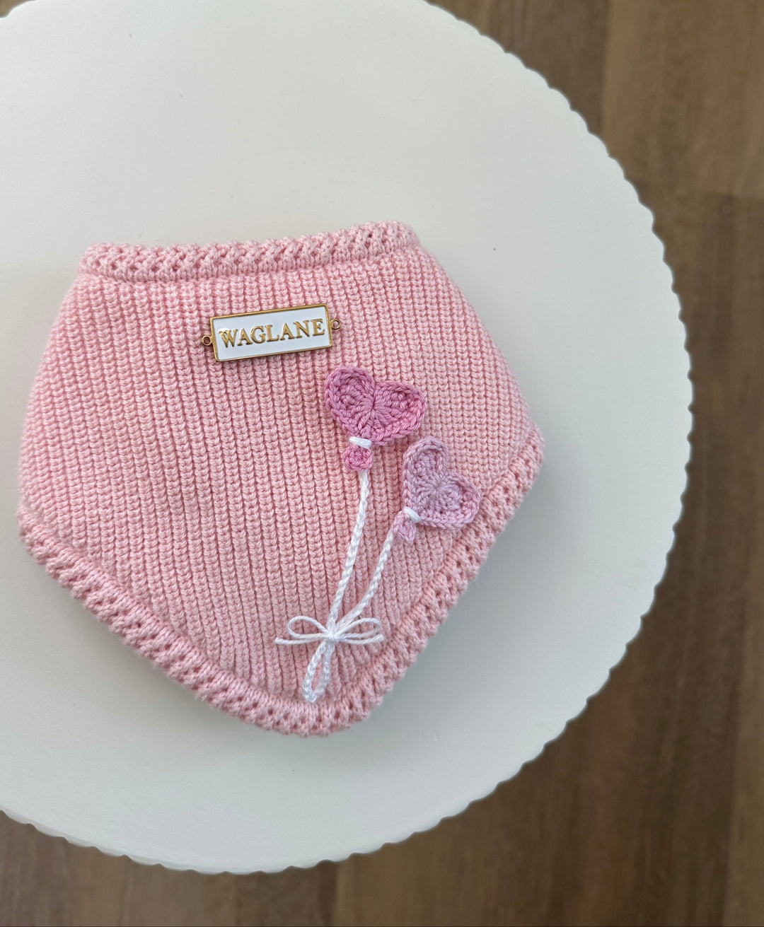Pink knit dog bandana with 2 crochet heart-shaped balloon appliqués in ose tones, tied with a white embroidered bow and finished with a white and gold rectangular Waglane™ tag on a round white platform.