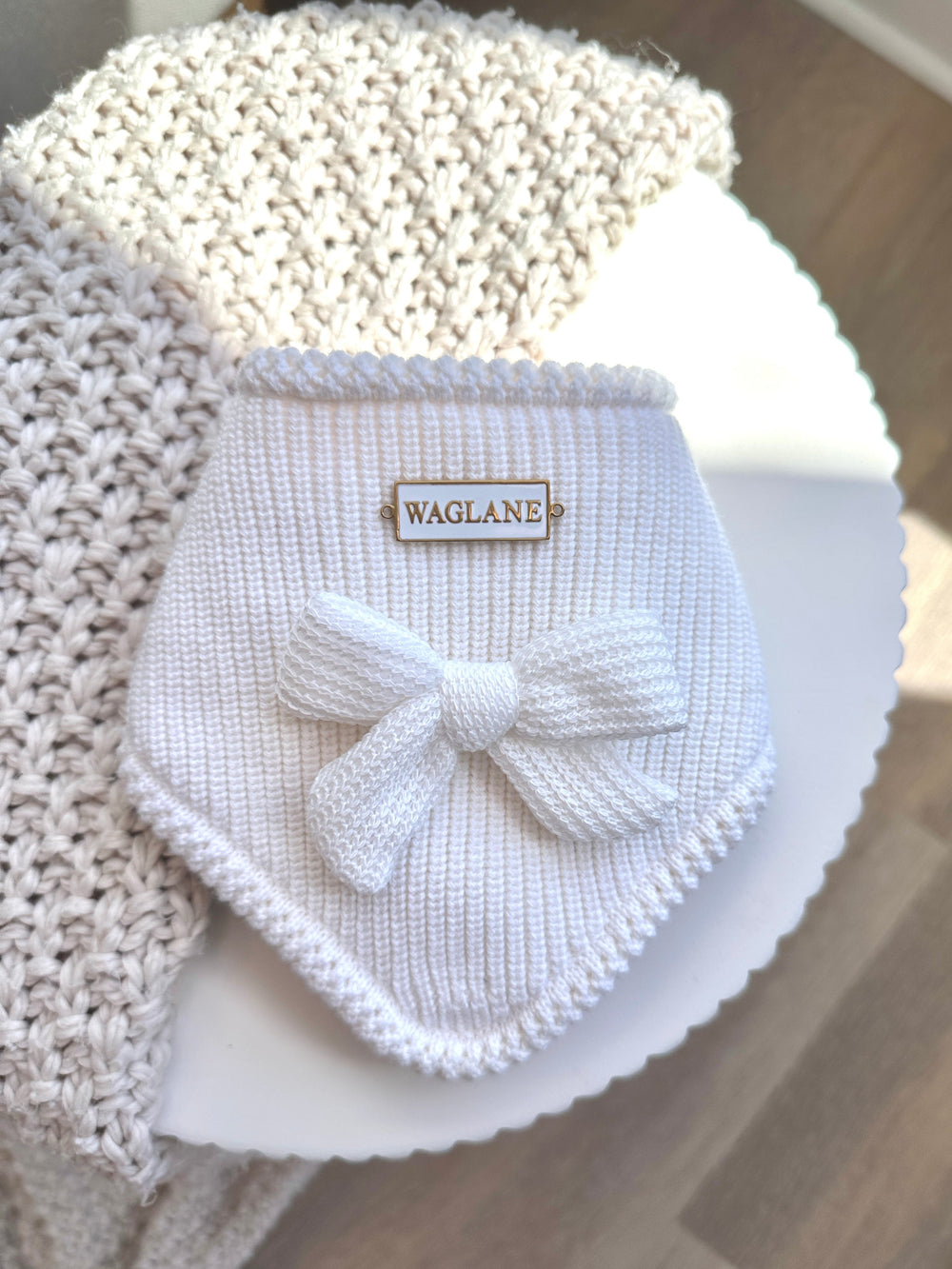 White knit pet bandana with white knitted large bow in center of bandana and 'WAGLANE' white metal rectangular label on a light plate background
