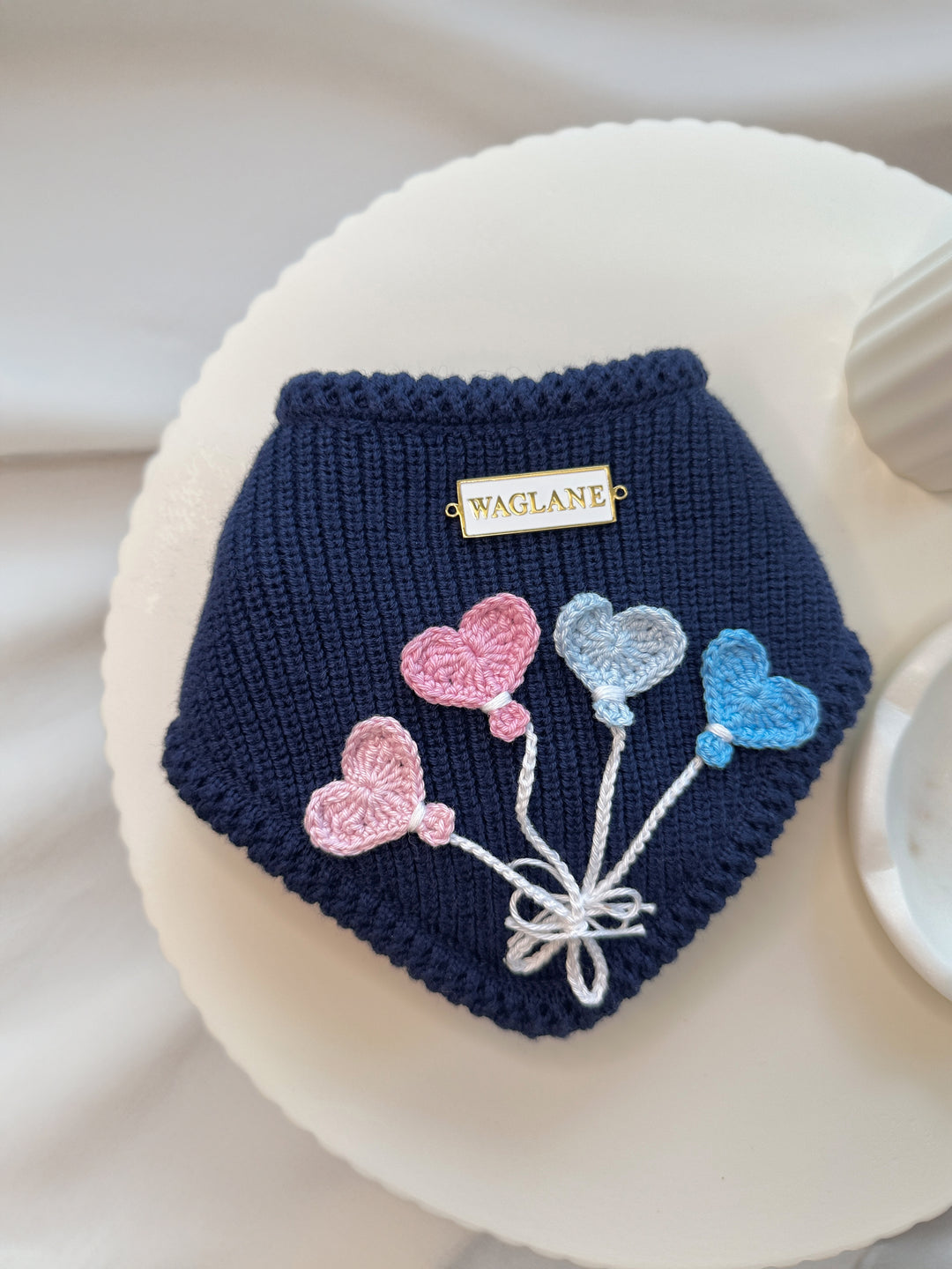 Navy blue knitted dog sweater with colorful crochet balloon appliques in shades of pink and light blue on a white round surface