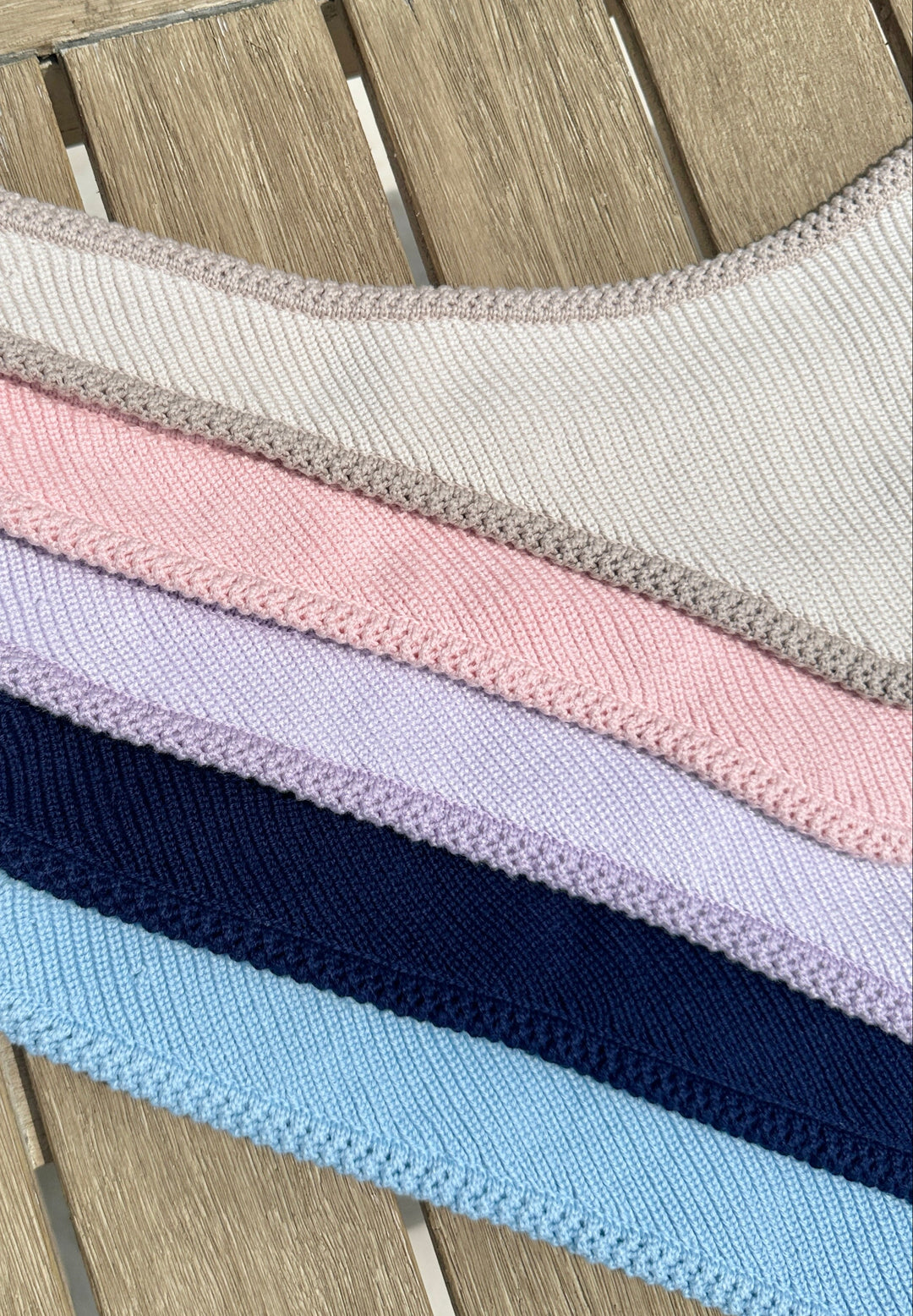 Stack of flat lay knit dog bandanas with luxe crochet edging, displayed on wood surface in multiple colors including two-tone tan, blush pink, two-tone lavender, navy blue, and light blue, showcasing personalized color options available for order