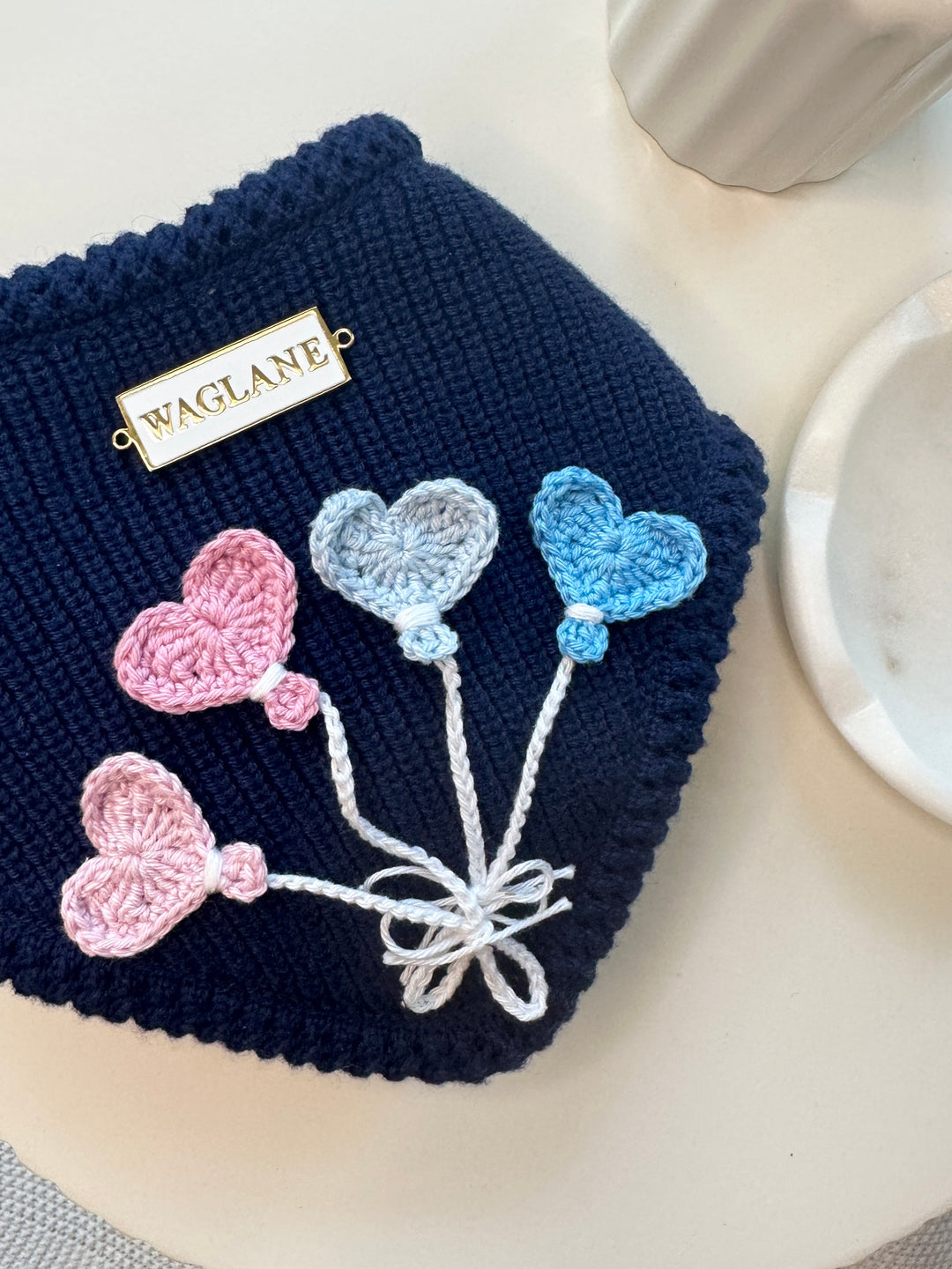 Navy blue hand-knit dog bandana with heart-shaped crochet balloon appliqués in pink, blue, and lavender tones, tied with white embroidered strings and finished with a white and gold rectangular WAGLANE™ logo tag, styled on a cream tabletop.